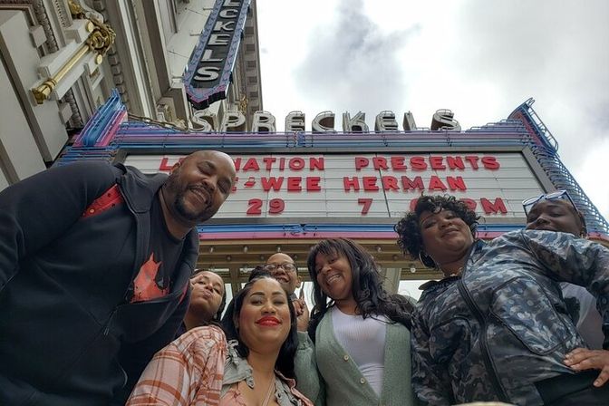 Group of smiling friends taking a low-angle selfie beneath a vintage downtown theater marquee with red letters and an overcast sky