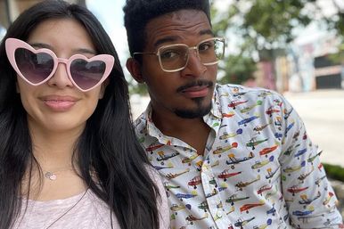 Outdoor selfie of two people on a sunny city sidewalk — woman with long dark hair wearing pink heart-shaped sunglasses and a small pendant, man wearing clear-framed glasses and a white button-down shirt covered in colorful vintage airplane prints.