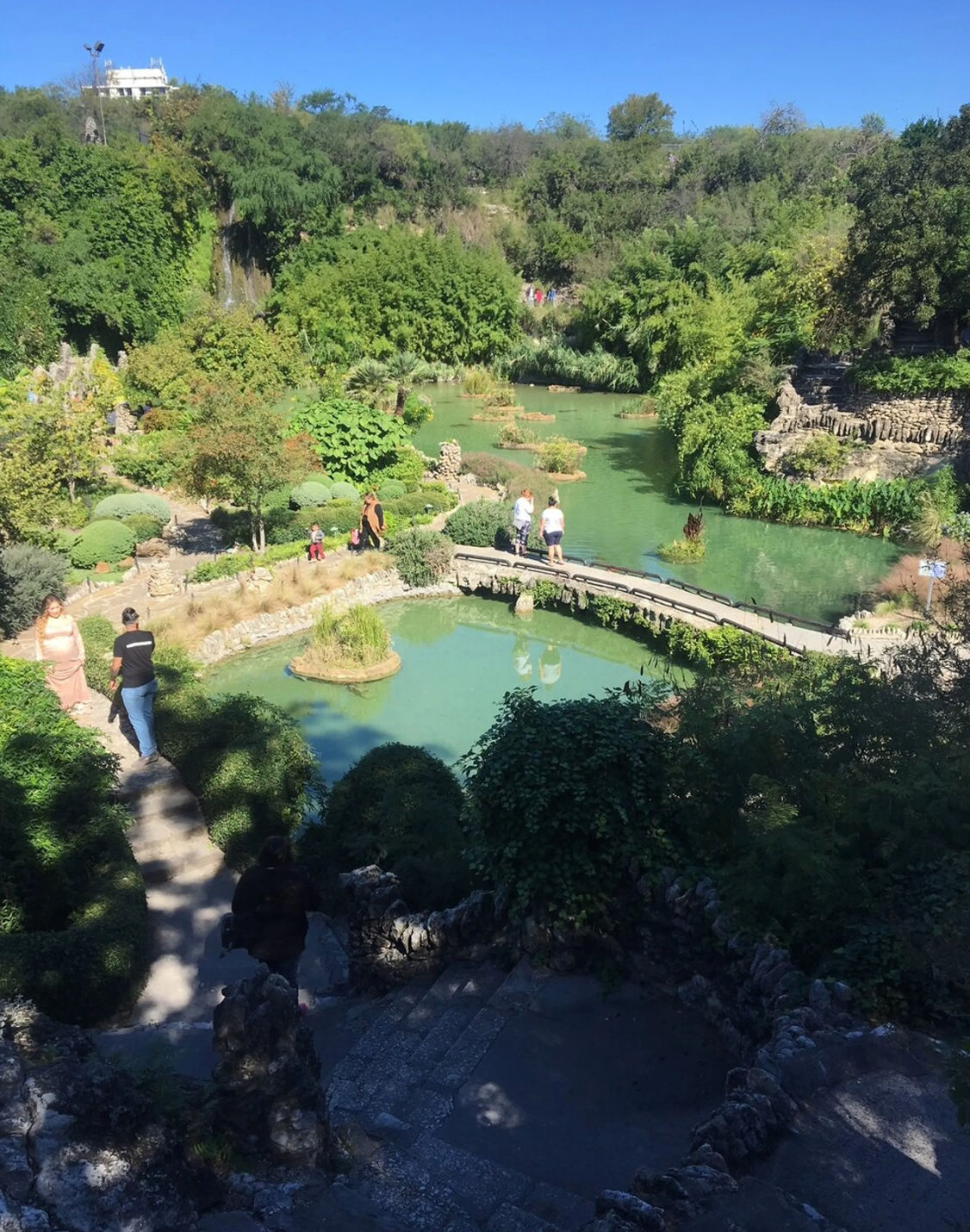 Sunlit hillside garden with turquoise ponds, small waterfall, arched stone footbridge, ornamental islands and people strolling manicured paths