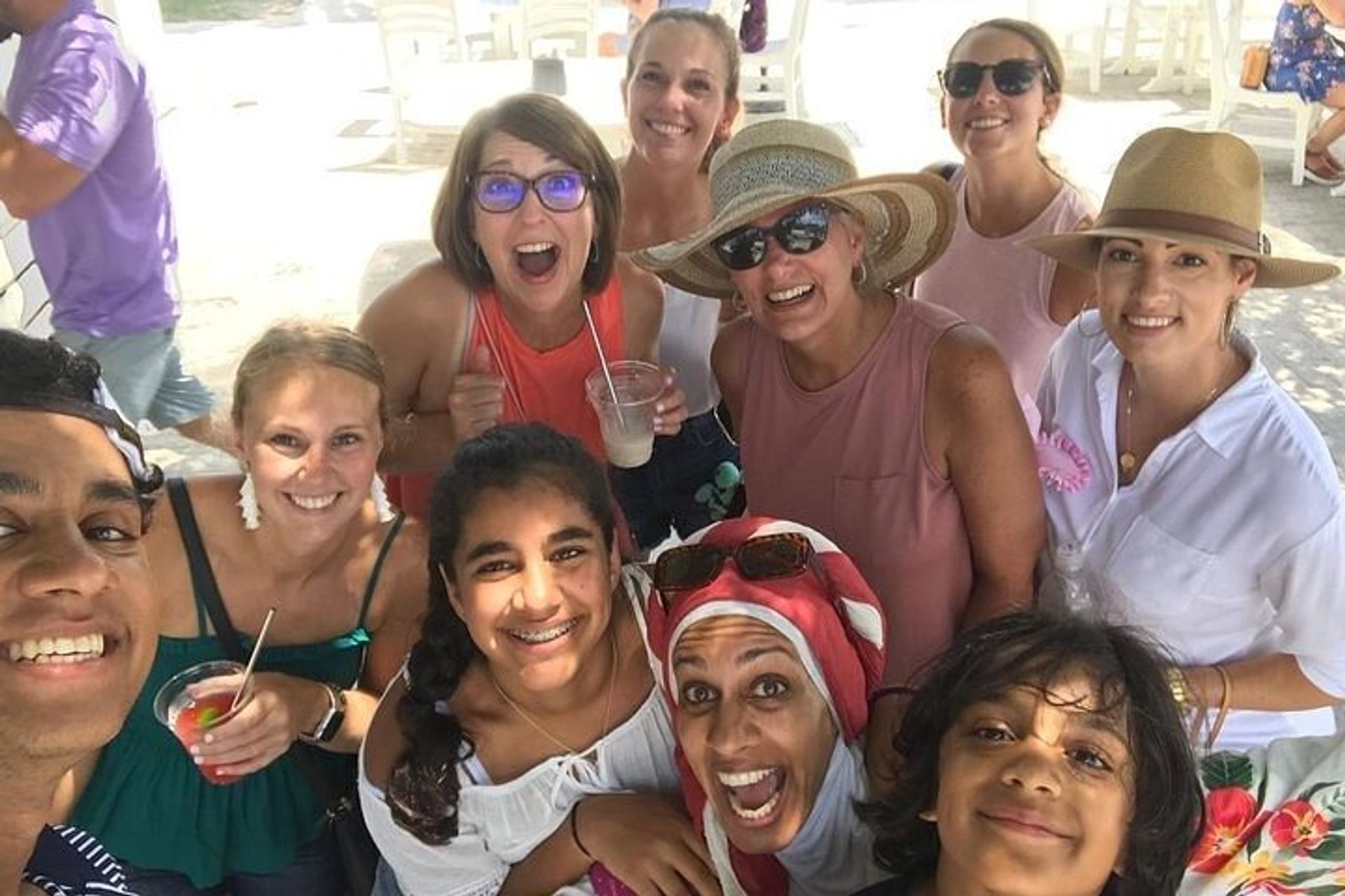 Cheerful multi-generational group selfie on a sunny beachside patio — friends and family in sun hats and sunglasses holding cold drinks.