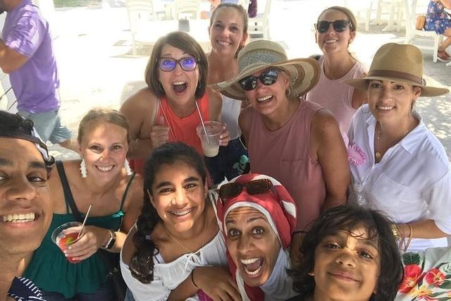 Cheerful multi-generational group selfie on a sunny beachside patio — friends and family in sun hats and sunglasses holding cold drinks.