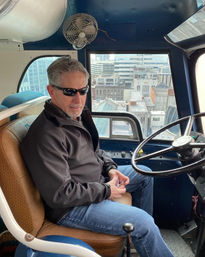 Person wearing sunglasses and a dark jacket seated in a bus cockpit at the wheel, hands folded, vintage fan overhead and downtown rooftops visible through the front window.