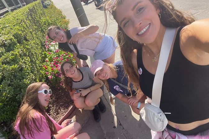 Group selfie of five smiling friends on a sunny sidewalk by low hedges and pink flowers, casually dressed in summer outfits including white sunglasses and a pink baseball cap.