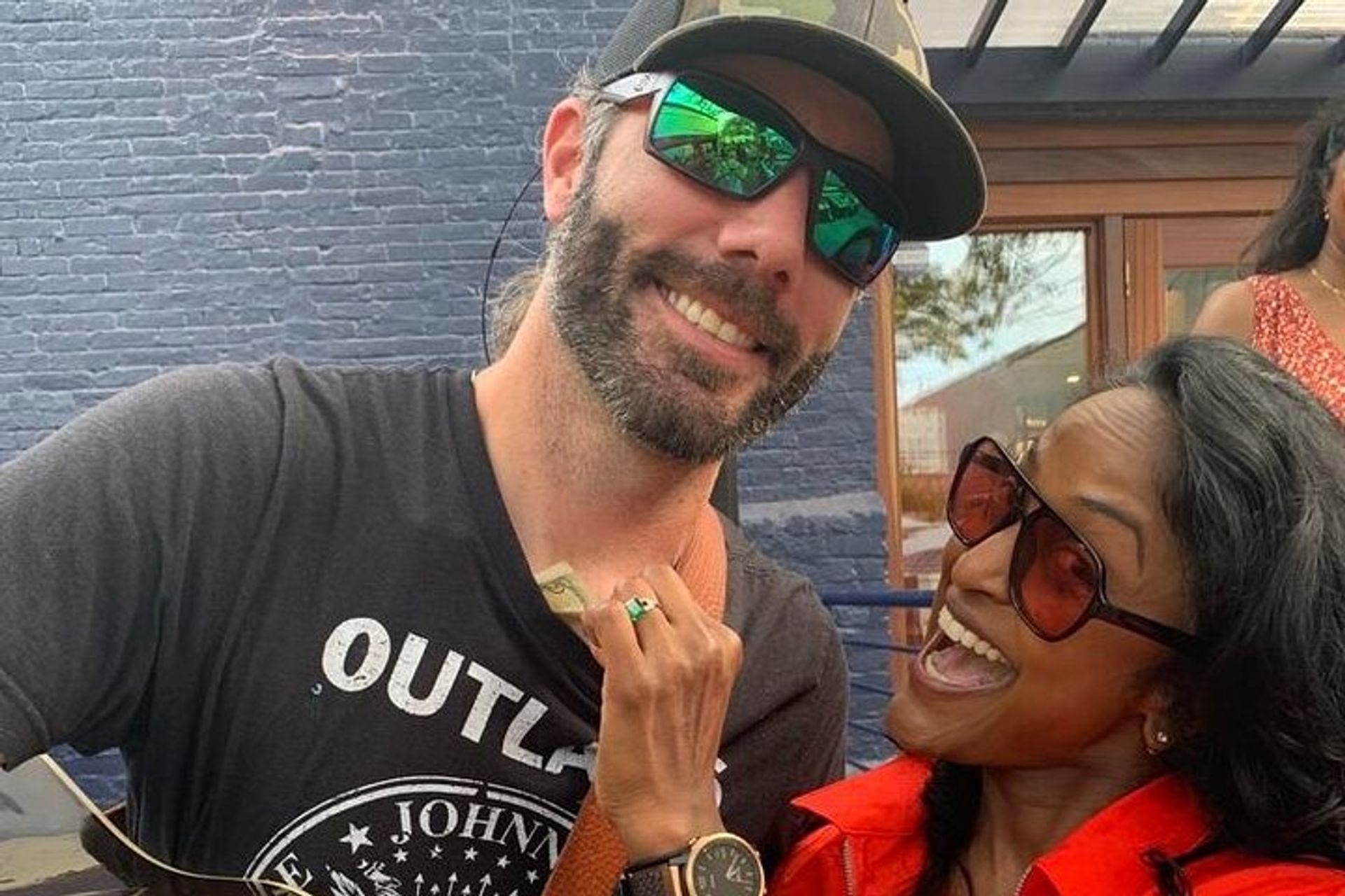 Two friends laughing on an outdoor patio — bearded man in a baseball cap and reflective sunglasses and a woman in a red jacket and oversized shades playfully holding cash near his chest, blue brick wall and window in the background.