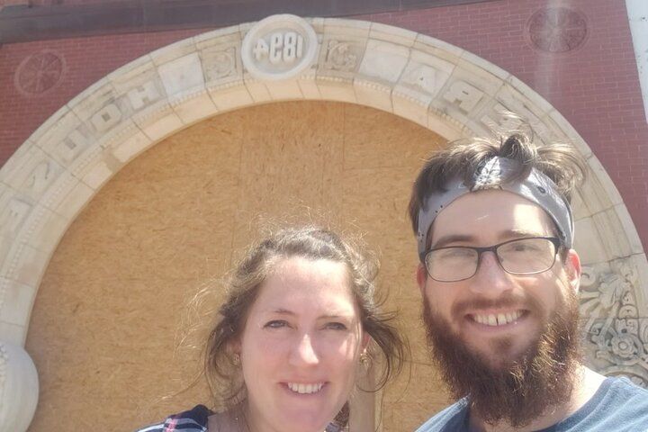 Smiling man with beard and headband and woman taking a sunny selfie in front of a boarded-up historic storefront with decorative stone arch.