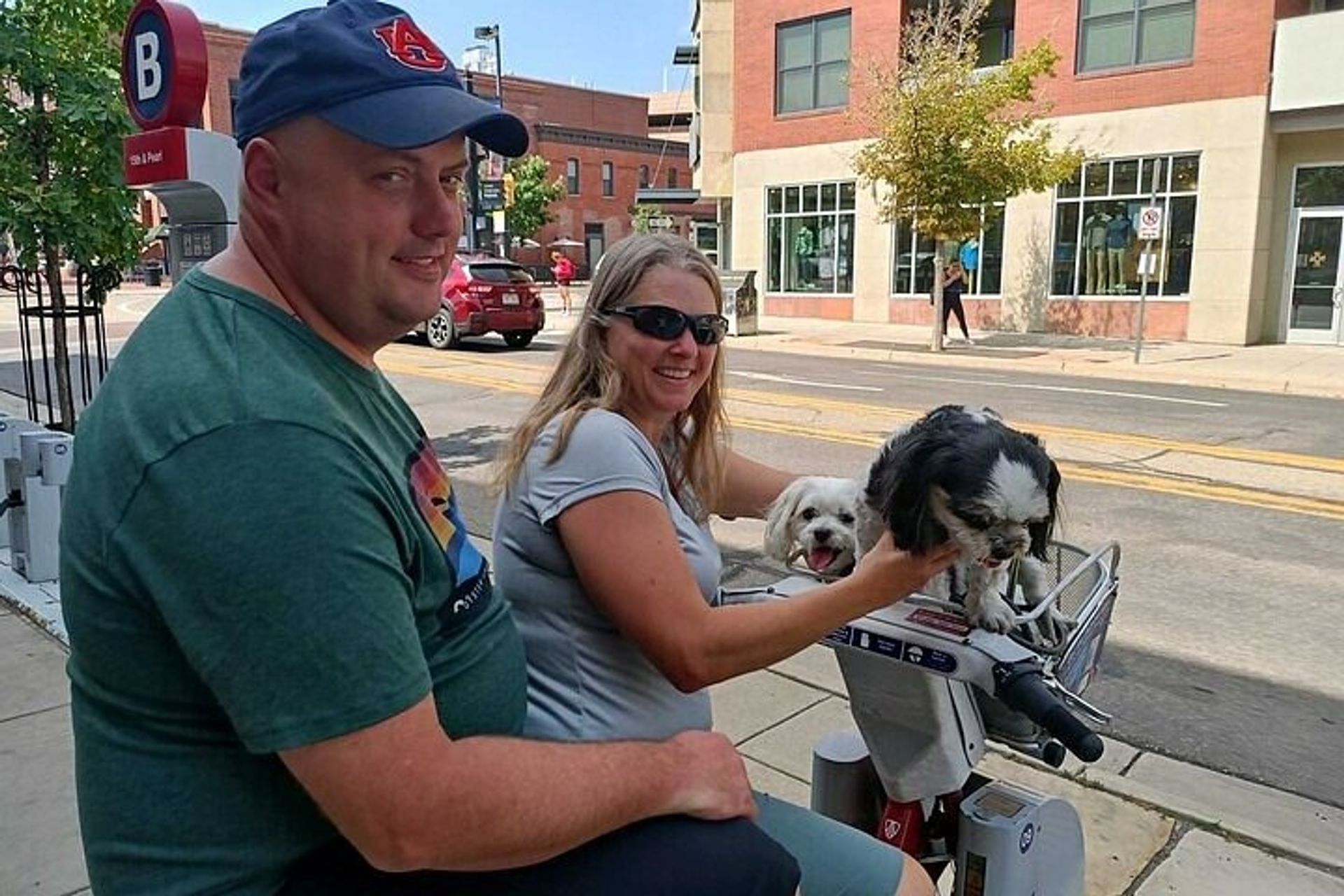 Smiling couple on a shared electric bike on a downtown city sidewalk, woman holding two small dogs — a black-and-white pup leaning from the front basket and a white pup peeking out, brick storefronts and a street in the background.
