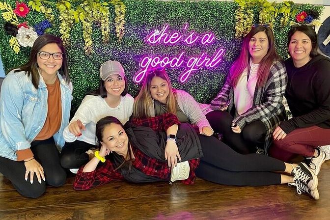 Six women in casual outfits pose and smile on a wooden floor in front of a green floral backdrop with a pink neon "she's a good girl" sign.