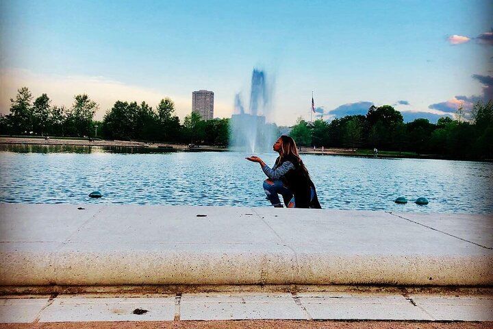 Person crouching at the edge of an urban park fountain, playfully blowing a kiss toward the central water jet with trees, a tall building and a flag on the skyline at dusk.