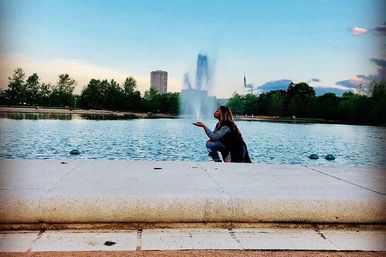Person crouching at the edge of an urban park fountain, playfully blowing a kiss toward the central water jet with trees, a tall building and a flag on the skyline at dusk.