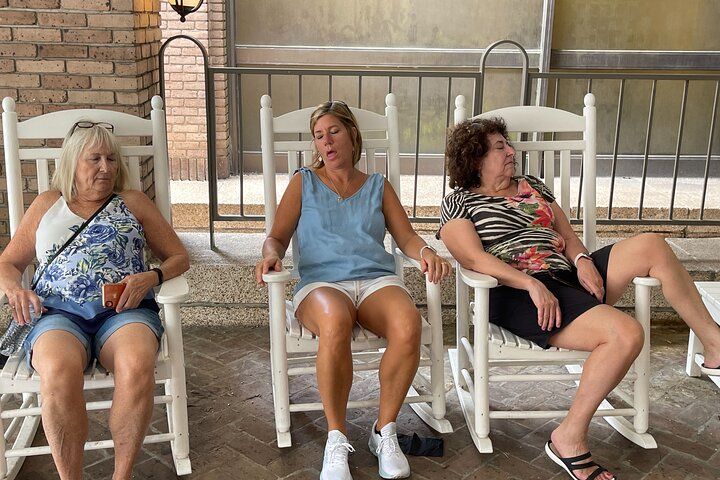 Three women relaxing in white wooden rocking chairs on a covered porch patio — casual summer scene with patterned tops, shorts, and sneakers, enjoying a slow, lazy moment.