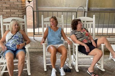 Three women relaxing in white wooden rocking chairs on a covered porch patio — casual summer scene with patterned tops, shorts, and sneakers, enjoying a slow, lazy moment.