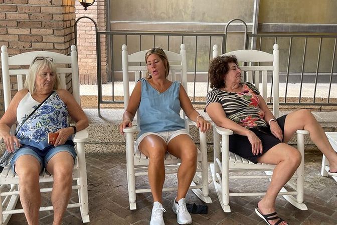Three women relaxing in white wooden rocking chairs on a covered porch patio — casual summer scene with patterned tops, shorts, and sneakers, enjoying a slow, lazy moment.