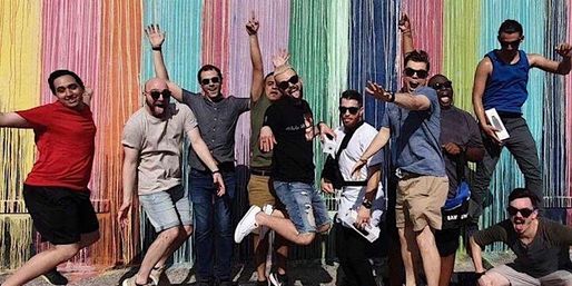 Group of friends jumping and posing in front of a vibrant rainbow string mural on an outdoor wall — playful street-art photo of people mid-air and smiling.