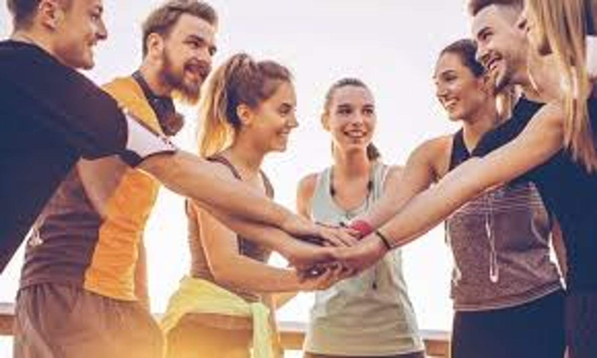 Group of young adults in athletic wear joining hands in an outdoor team workout huddle at sunset — energetic group fitness motivation.