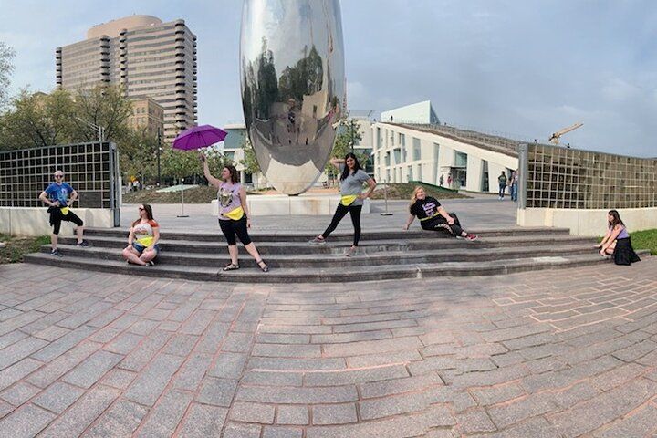 Six people spaced on stone steps in an urban plaza posing around a large mirrored egg-shaped public sculpture, one holding a purple umbrella, with modern buildings and a paved forecourt in the background.