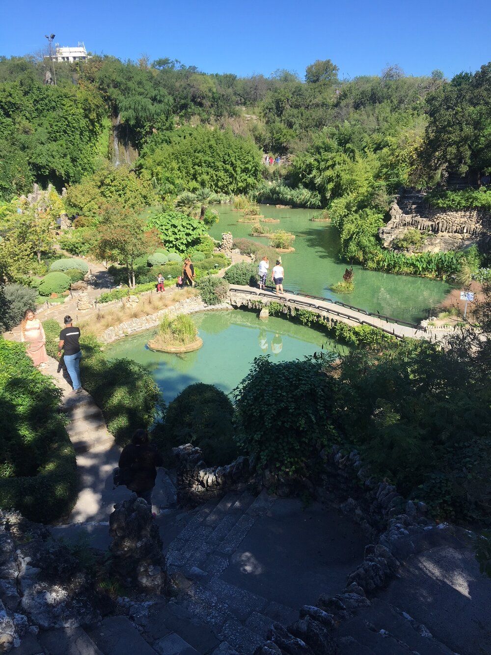 Serene hillside water garden with turquoise pond, small curved stone bridge, island plantings and winding stone steps amid lush shrubs and trees, visitors strolling on a sunny day.