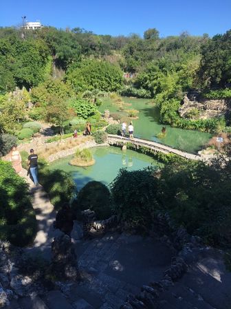 Serene hillside water garden with turquoise pond, small curved stone bridge, island plantings and winding stone steps amid lush shrubs and trees, visitors strolling on a sunny day.