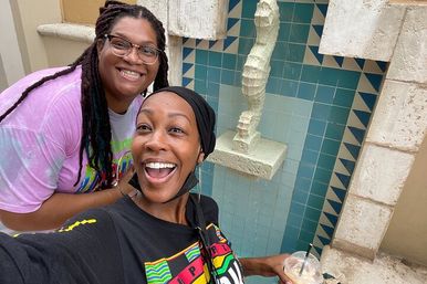 Two smiling friends take a selfie in front of a turquoise-tiled fountain niche with a textured seahorse sculpture; one holds an iced coffee.