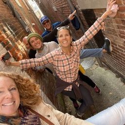 Five friends smiling for a selfie, playfully balancing with arms out in a narrow brick alleyway in an urban downtown setting, wearing casual fall clothes and beanies.