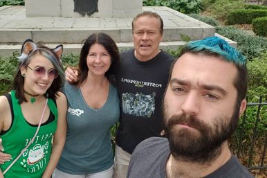 Group selfie of four adults in a city park in front of a stone monument — smiling woman with cat-ear headband and green tank top, woman in a gray V-neck tee, man in a black graphic shirt, and a bearded man with blue-dyed hair taking the photo.