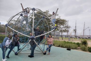 Group of friends climbing a spherical metal-and-rope playground structure at a waterfront park playground, with a promenade, tall ship masts and overcast sky in the background.