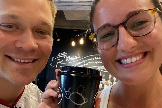 Selfie of two smiling people in a cafe holding a black takeaway coffee cup with a white doodle, warm hanging lights and a chalkboard wall in the background.