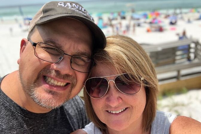 Smiling couple selfie on a sunny sandy beach, one wearing sunglasses and the other a cap, with a crowded shoreline and wooden boardwalk in the background