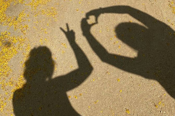 Sunlit concrete pavement sprinkled with yellow flower petals and playful shadows of two people — one flashing a peace sign, the other making a heart with their hands.