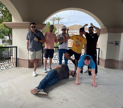 Seven friends posing playfully under an arched outdoor gazebo with palm trees and rocky hills visible through the openings — two men on the ground (one lying down, one doing a wheelbarrow) and the others standing, flexing and laughing in casual summer clothes and sunglasses at a sunny plaza.