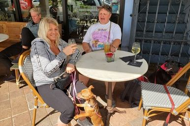 Sunlit sidewalk café patio with a smiling woman holding a glass of white wine at a round bistro table, a man with a beer, a small brown dog on her lap and a black dog nearby on a leash.