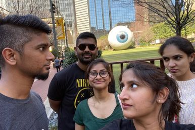 Playful group selfie of five young adults in a downtown urban plaza, posing with varied expressions in front of a giant eyeball sculpture on a grassy lawn with trees and glass skyscrapers in the background.