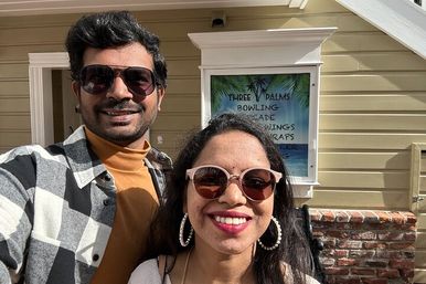 Sunny selfie of two people wearing sunglasses — man in a checkered jacket and mustard turtleneck and woman with pink-framed shades and hoop earrings — standing outside a beachside storefront with a palm-themed poster and brick ledge.