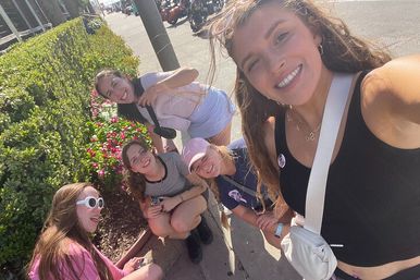 Group of five friends smiling for a sunny sidewalk selfie by a bright pink flower bed and trimmed hedge, casual summer outfits and crossbody bags with parked motorcycles and a city street in the background.