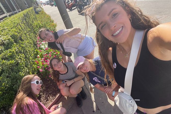 Group of five friends smiling for a sunny sidewalk selfie by a bright pink flower bed and trimmed hedge, casual summer outfits and crossbody bags with parked motorcycles and a city street in the background.
