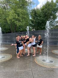Seven women in matching black-and-white outfits barefoot, laughing and posing as vertical water jets spray around them at an outdoor splash pad in a tree-lined park with an engraved stone wall behind them.