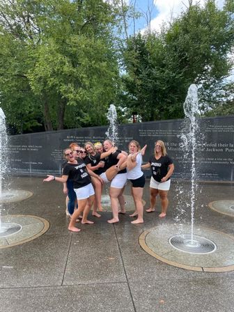 Seven women in matching black-and-white outfits barefoot, laughing and posing as vertical water jets spray around them at an outdoor splash pad in a tree-lined park with an engraved stone wall behind them.