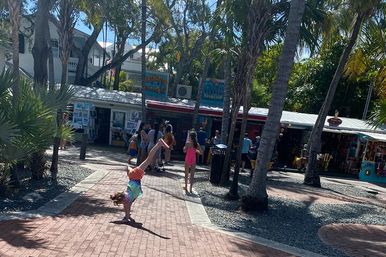 Child doing a cartwheel on a brick walkway in a palm-lined, beachside outdoor market plaza with tourists and colorful souvenir shops under sunny skies