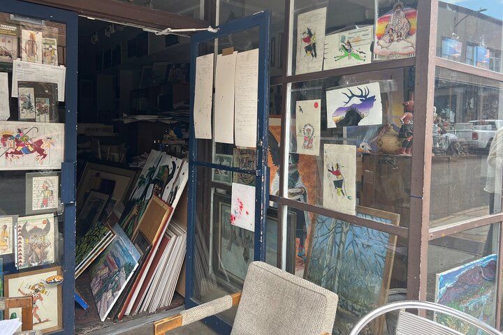 Cozy art shop storefront with open door revealing stacked framed paintings and colorful prints in the window, sidewalk chair and street reflections