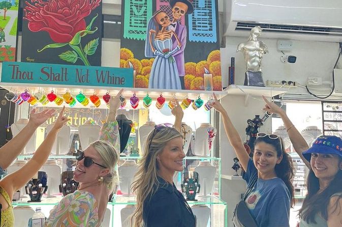 Group of friends inside a colorful boutique gift shop pointing at a teal sign reading 'Thou Shalt Not Whine' above a Day of the Dead skeleton couple painting, heart-shaped string lights and glass jewelry displays.