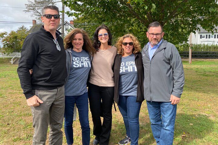 Five adults posing on a suburban lawn under a leafy tree, wearing casual jackets and jeans; two women in matching "Do Epic Sh*t" graphic tees — relaxed outdoor group photo.