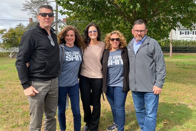 Five adults posing on a suburban lawn under a leafy tree, wearing casual jackets and jeans; two women in matching "Do Epic Sh*t" graphic tees — relaxed outdoor group photo.