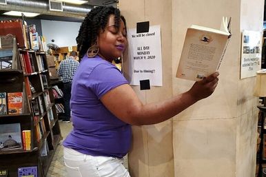 Woman in a purple shirt reading a paperback while leaning against a pillar inside a cozy independent bookstore with tall shelves of books in the background