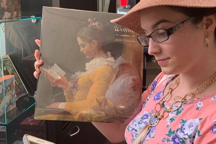 Person in floral dress, glasses and sunhat holds a 2020 calendar featuring a classical painting of a seated woman reading, in a shop display.