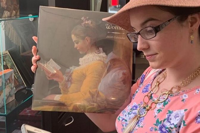 Person in floral dress, glasses and sunhat holds a 2020 calendar featuring a classical painting of a seated woman reading, in a shop display.