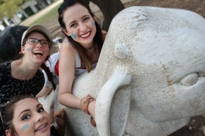 Three smiling friends with blue face paint take a playful selfie while leaning on a weathered white stone elephant sculpture in an outdoor park