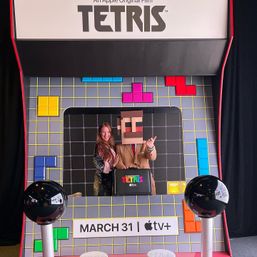Playful photo of two people in a giant Tetris-themed booth: one wearing a pixelated box head, colorful Tetrimino pieces on a gridded backdrop, and signage announcing the Tetris film with a March 31 streaming release.