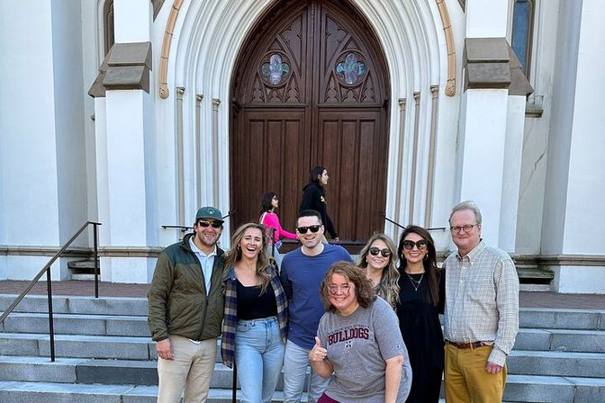 Smiling group of friends posing on stone steps in front of a gothic arched wooden church entrance; casual outfits and sunglasses, one person gives a thumbs-up, two pedestrians walk up the steps in the background.