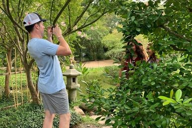 Man in a baseball cap photographing two smiling women peeking through leafy bushes in a tranquil Japanese-style garden with a stone lantern and pond
