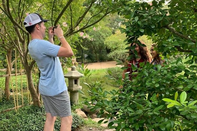Man in a baseball cap photographing two smiling women peeking through leafy bushes in a tranquil Japanese-style garden with a stone lantern and pond
