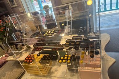 Acrylic display case of colorful artisanal chocolate bonbons and truffles arranged on black trays on a marble countertop by a sunlit shop window with a glimpse of a wooden boardwalk outside.